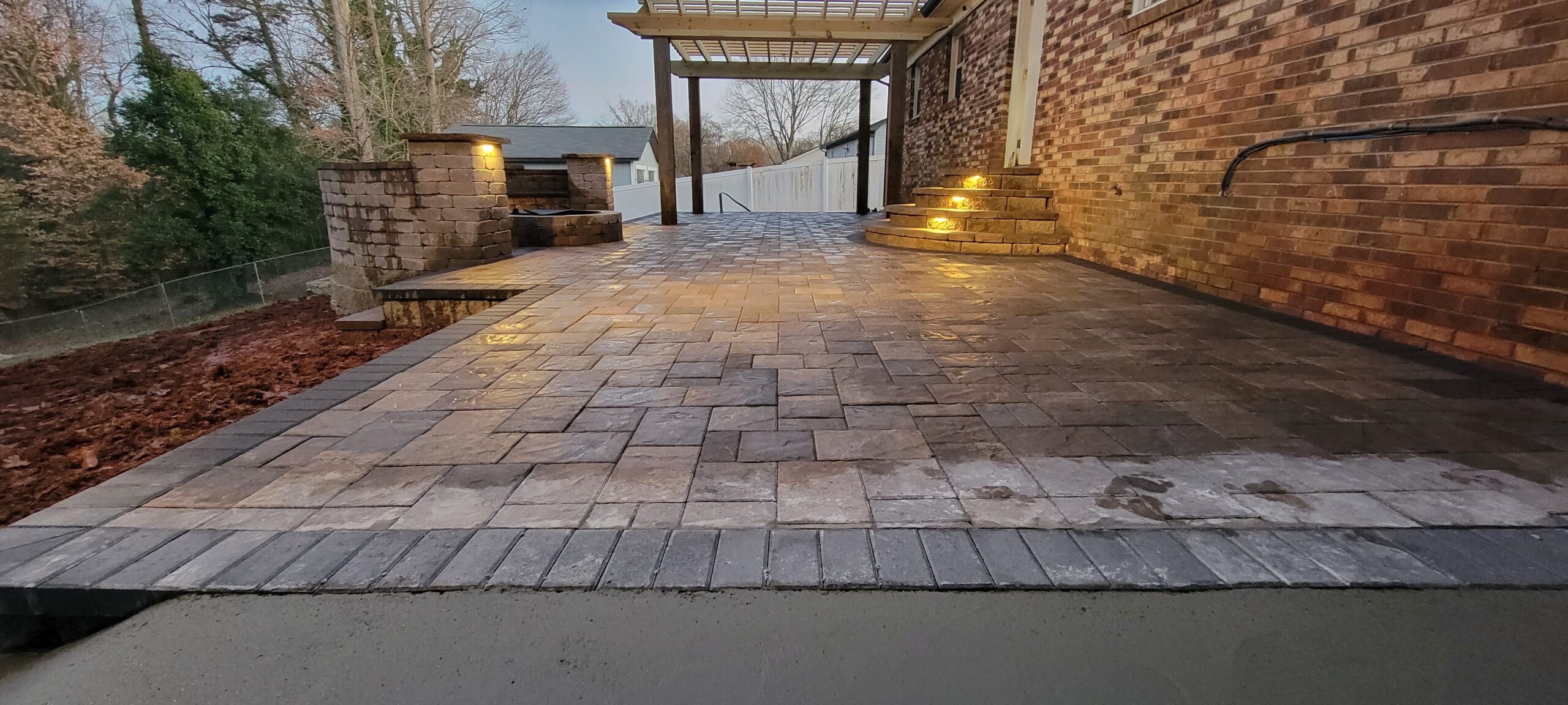 Paver patio with a dark border, curved steps and an open wood overhead structure beside a brick house