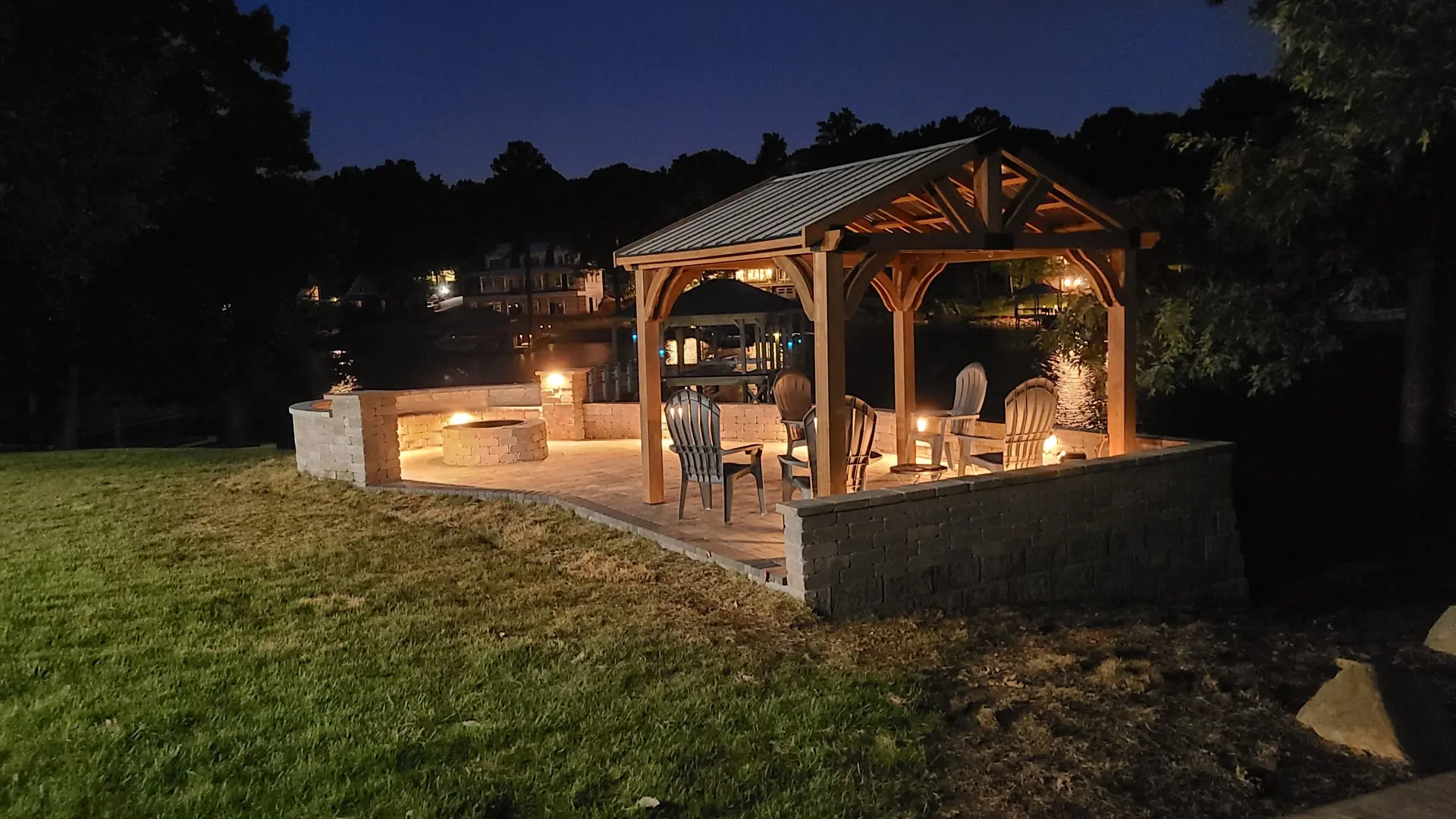 Illuminated lakeside patio with a wood pavilion, Adirondack chairs, round fire pit and seating walls at night